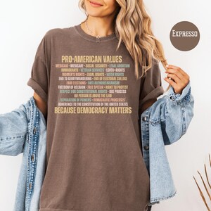Pode incluir: Uma camiseta marrom com o texto "PRO-AMERICAN VALUES" e uma lista de valores em v&aacute;rias cores. A frase "BECAUSE DEMOCRACY MATTERS" est&aacute; na parte inferior. A camiseta &eacute; usada com uma jaqueta jeans.