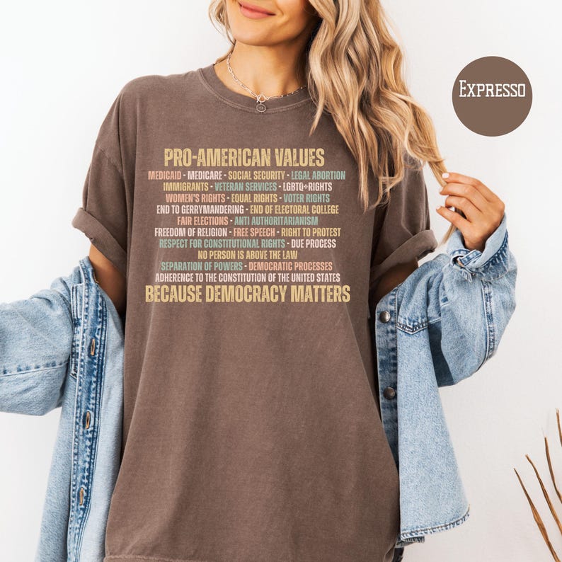 Pode incluir: Uma camiseta marrom com o texto "PRO-AMERICAN VALUES" e uma lista de valores em v&aacute;rias cores. A frase "BECAUSE DEMOCRACY MATTERS" est&aacute; na parte inferior. A camiseta &eacute; usada com uma jaqueta jeans.
