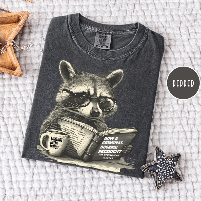 May include: Pepper-colored t-shirt featuring a raccoon wearing glasses and reading a book titled "How a Criminal Became President." The shirt also displays the text "Foxtrot Delta Tango" on a mug. The shirt is made of a soft material.