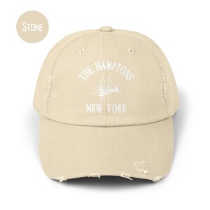 May include: A stone-colored distressed baseball cap with white embroidery that reads "THE HAMPTONS NEW YORK" and features a sailboat graphic.
