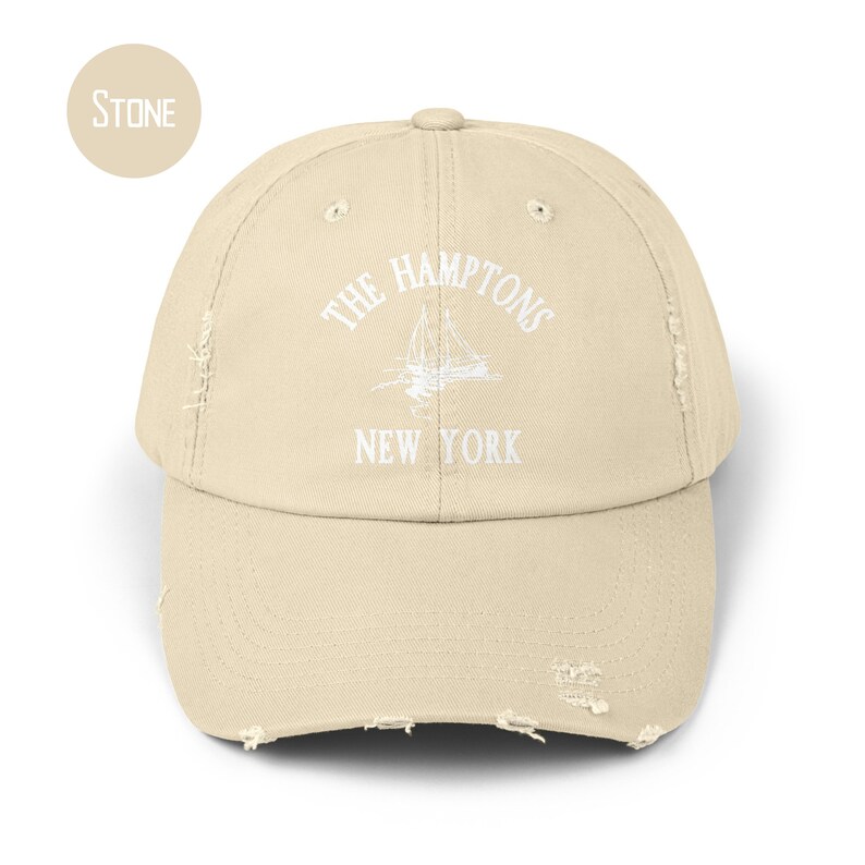 May include: A stone-colored distressed baseball cap with white embroidery that reads "THE HAMPTONS NEW YORK" and features a sailboat graphic.
