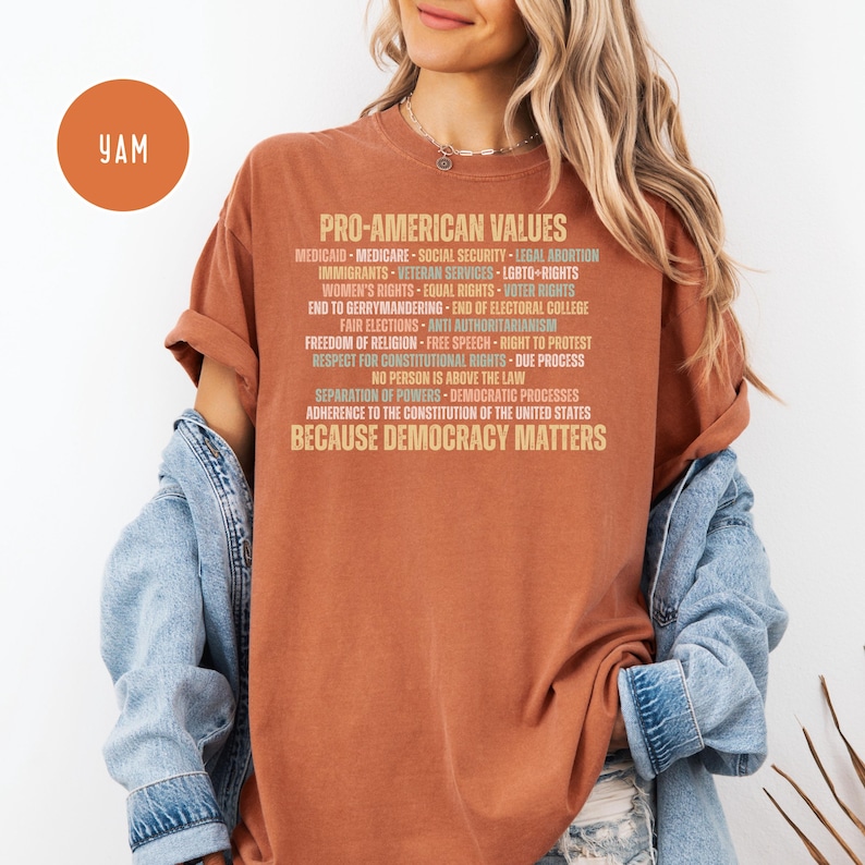 Pode incluir: Uma camiseta cor de ferrugem com o texto "PRO-AMERICAN VALUES" e uma lista de valores associados em uma fonte marrom clara. A frase "BECAUSE DEMOCRACY MATTERS" est&aacute; na parte inferior. A camiseta &eacute; usada por uma pessoa.