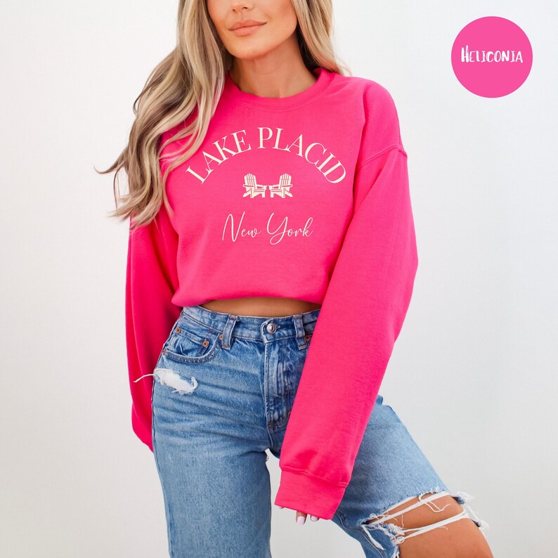 May include: A bright pink sweatshirt with the words "Lake Placid New York" in white script. The design includes two white Adirondack chairs. The sweatshirt is paired with blue jeans.