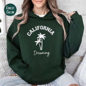 Puede incluir: Sudadera con capucha verde bosque con texto blanco que dice "CALIFORNIA DREAMING" y un gr&aacute;fico de dos palmeras.