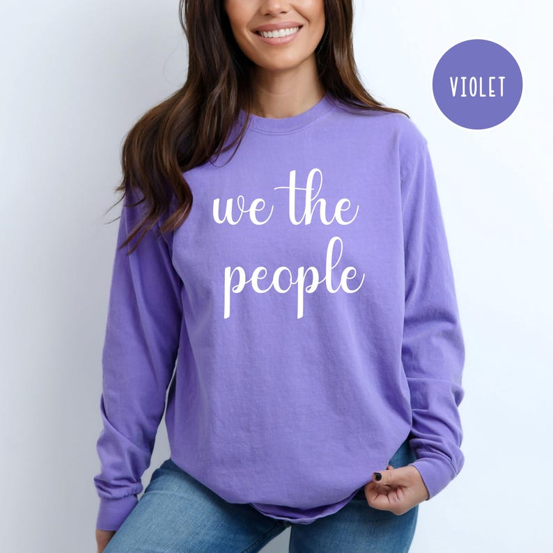 Puede incluir: Una camiseta de manga larga de color morado con el texto "we the people" en letras blancas.