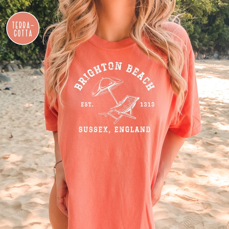 K&ouml;nnte beinhalten: Ein korallenfarbenes T-Shirt mit einem wei&szlig;en Grafikdesign, auf dem "Brighton Beach Est. 1313 Sussex, England" mit einem Sonnenschirm und einem Liegestuhl steht.