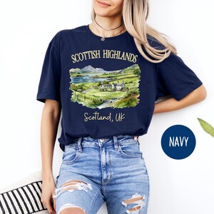 Puede incluir: Camiseta azul marino con una ilustraci&oacute;n en acuarela de las Tierras Altas escocesas. El dise&ntilde;o incluye un paisaje con monta&ntilde;as, un lago y edificios, con el texto "Scottish Highlands" y "Scotland, UK".