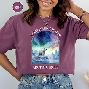 Op de afbeelding: Bessenkleurig T-shirt met een afbeelding van een ijsbeer op een ijsberg onder het noorderlicht. Het ontwerp bevat de tekst "NORTHERN LIGHTS" en "ARCTIC CIRCLE" in het wit. Het shirt is gemaakt van een zacht materiaal.