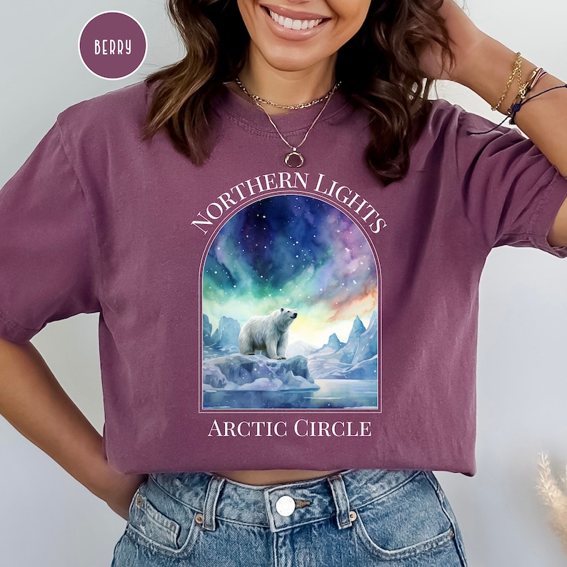 Op de afbeelding: Bessenkleurig T-shirt met een afbeelding van een ijsbeer op een ijsberg onder het noorderlicht. Het ontwerp bevat de tekst "NORTHERN LIGHTS" en "ARCTIC CIRCLE" in het wit. Het shirt is gemaakt van een zacht materiaal.
