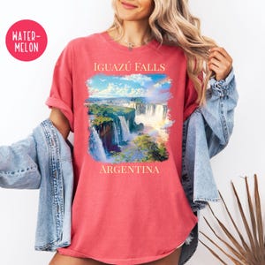 Pu&ograve; includere: T-shirt color anguria con un'immagine panoramica delle cascate dell'Iguaz&uacute; in Argentina. La grafica include il testo "IGUAZ&Uacute; FALLS" sopra le cascate e "ARGENTINA" sotto. La maglietta ha maniche arrotolate e una vestibilit&agrave; rilassata.