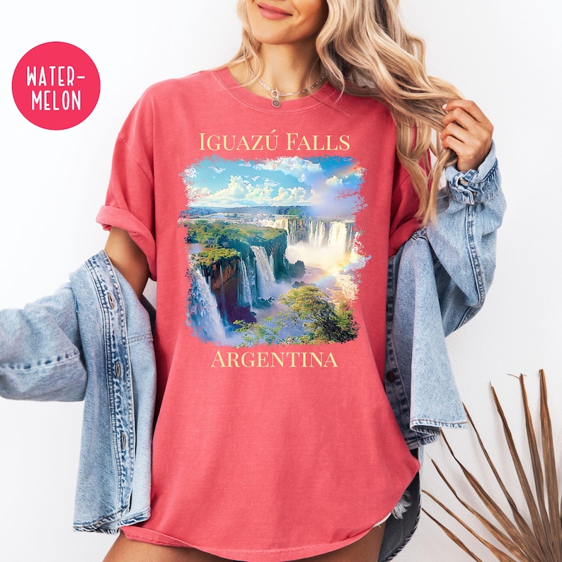 Pu&ograve; includere: T-shirt color anguria con un'immagine panoramica delle cascate dell'Iguaz&uacute; in Argentina. La grafica include il testo "IGUAZ&Uacute; FALLS" sopra le cascate e "ARGENTINA" sotto. La maglietta ha maniche arrotolate e una vestibilit&agrave; rilassata.