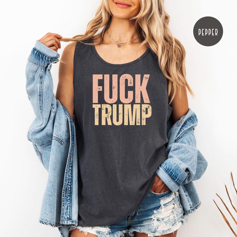 K&ouml;nnte beinhalten: Dunkelgraues Tanktop mit dem Text "FUCK TRUMP" in Rosa und Gelb. Das Tanktop wird mit einer hellblauen Jeansjacke getragen. Das Wort "PEPPER" befindet sich in einem schwarzen Kreis oben rechts.