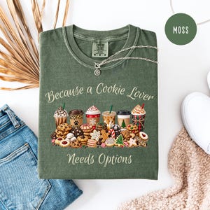 May include: A moss green t-shirt featuring a graphic print of various coffee cups and assorted cookies. The text "Because a Cookie Lover Needs Options" is printed in a light cream color above and below the illustration of the treats.