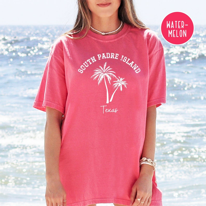 May include: A pink t-shirt with the text "South Padre Island Texas" and two palm trees in white. The shirt is a relaxed fit and has a crew neck.
