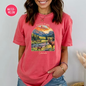 May include: A red t-shirt featuring a vintage-style graphic of a mountain scene with a river and elk. The graphic reads "Estes Park Colorado" in a white font.
