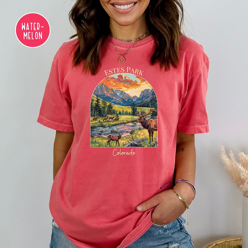 May include: A red t-shirt featuring a vintage-style graphic of a mountain scene with a river and elk. The graphic reads "Estes Park Colorado" in a white font.