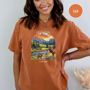May include: A rust-coloured t-shirt with a graphic print of a mountain scene with a river and elk. The text "ESTES PARK Colorado" is printed on the shirt.