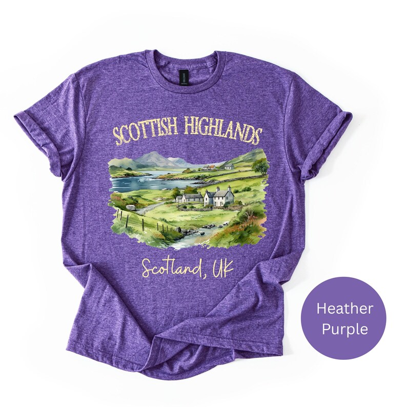 Puede incluir: Camiseta morada jaspeada con una ilustraci&oacute;n en acuarela de las Tierras Altas escocesas. El dise&ntilde;o incluye un paisaje con un lago, monta&ntilde;as y edificios, con el texto "Scottish Highlands" y "Scotland, UK".