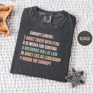 May include: A dark gray t-shirt with the text "CORRUPT LEADERS: TARGET TRUTH WITH FEAR, RIG MEDIA FOR CONTROL, UNDERMINE RULE OF LAW, MARKET LIES AS LEADERSHIP, PARDON THE CORRUPT" in various colors. The shirt is labeled "Pepper".