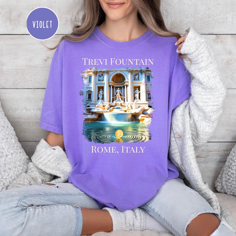May include: A violet t-shirt with a graphic of the Trevi Fountain in Rome, Italy. The image depicts the fountain's architecture and water features. The text "TREVI FOUNTAIN" and "ROME, ITALY" are printed above and below the image.