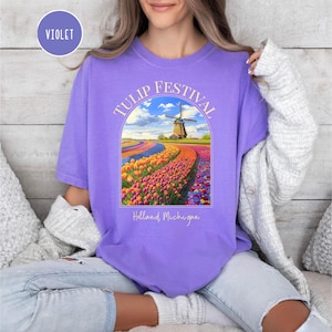 May include: A purple t-shirt with the text "TULIP FESTIVAL" in a pink arch above a colorful image of a field of tulips and a windmill. The text "Holland, Michigan" is below the image.