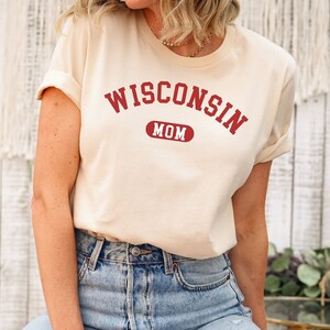 Puede incluir: Una camiseta de color crema con el texto "WISCONSIN MOM" en letras rojas. El texto est&aacute; en una fuente de estilo universitario.