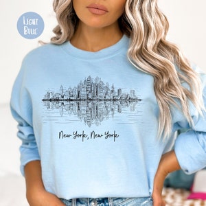 K&ouml;nnte beinhalten: Ein hellblaues Sweatshirt mit einer schwarz-wei&szlig;en Grafik der Skyline von New York City, die sich im Wasser spiegelt. Der Text "New York, New York" ist unter der Grafik aufgedruckt.