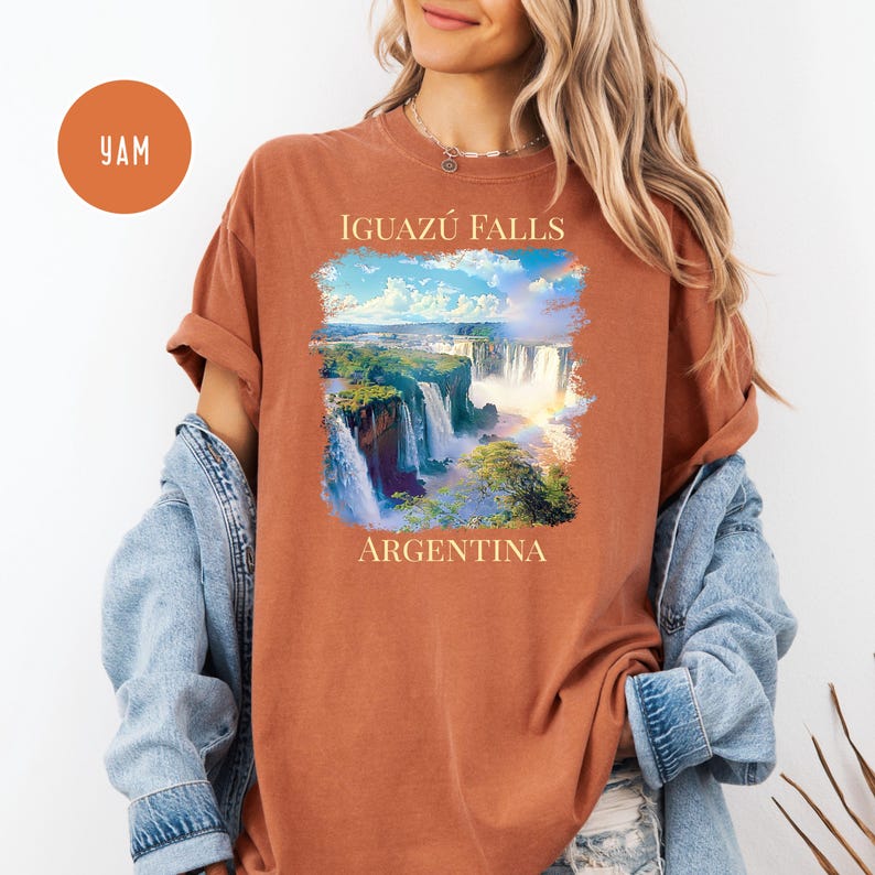 Pu&ograve; includere: T-shirt color ruggine con un'illustrazione panoramica delle cascate dell'Iguaz&uacute; in Argentina. Il design include il testo "IGUAZU FALLS" sopra l'immagine e "ARGENTINA" sotto. La maglietta ha maniche corte e una vestibilit&agrave; rilassata.