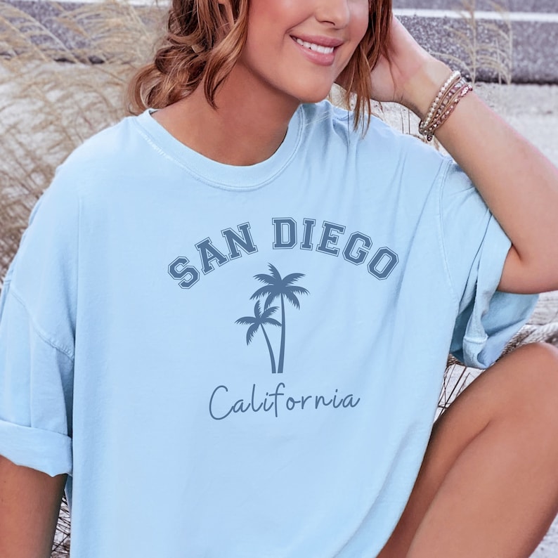 Może przedstawiać: Jasnoniebieska koszulka z nadrukiem graficznym palm i tekstem "SAN DIEGO California".
