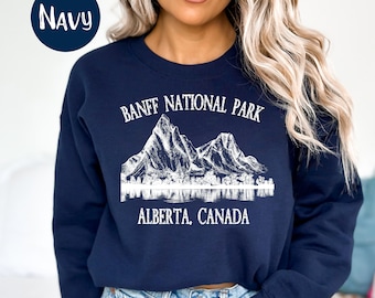 Puede incluir: Sudadera azul marino con un gráfico blanco de montañas y un lago, con el texto "BANFF NATIONAL PARK ALBERTA, CANADA". La palabra "NAVY" está en un círculo azul marino en la esquina superior izquierda. La sudadera es de cuello redondo.