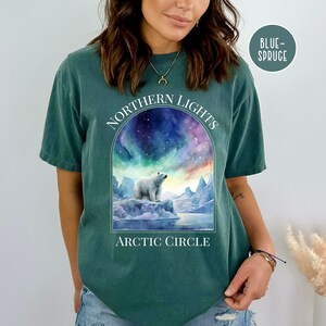 Op de afbeelding: Een tealkleurig T-shirt met een afbeelding van een ijsbeer op een ijsberg onder het noorderlicht. De tekst "NORTHERN LIGHTS" staat boven de afbeelding, met "ARCTIC CIRCLE" eronder. Het shirt is gelabeld "BLUE-SPRUCE".