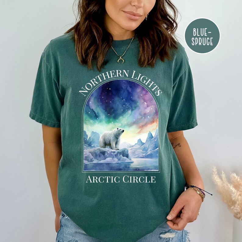 Op de afbeelding: Een tealkleurig T-shirt met een afbeelding van een ijsbeer op een ijsberg onder het noorderlicht. De tekst "NORTHERN LIGHTS" staat boven de afbeelding, met "ARCTIC CIRCLE" eronder. Het shirt is gelabeld "BLUE-SPRUCE".