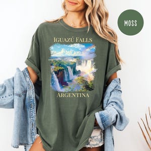 Pu&ograve; includere: T-shirt verde muschio con un'immagine delle cascate dell'Iguaz&ugrave; in Argentina. Il design include il testo "IGUAZ&Uacute; FALLS" sopra l'immagine della cascata e "ARGENTINA" sotto. Un'etichetta circolare "MOSS" &egrave; nell'angolo in alto a destra.