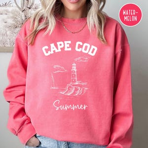 Könnte beinhalten: Ein rosafarbenes Sweatshirt mit einem weißen Grafikdesign, auf dem "Cape Cod Summer" steht und ein Leuchtfeuer und ein Segelboot abgebildet sind. Das Sweatshirt hat eine lockere Passform und einen Rundhalsausschnitt.