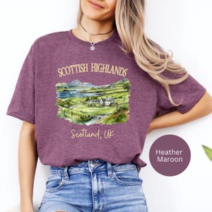 Puede incluir: Camiseta color granate con una ilustraci&oacute;n en acuarela de las Tierras Altas escocesas. El dise&ntilde;o incluye un paisaje con monta&ntilde;as, un lago y casas, con el texto "Scottish Highlands" y "Scotland, UK".