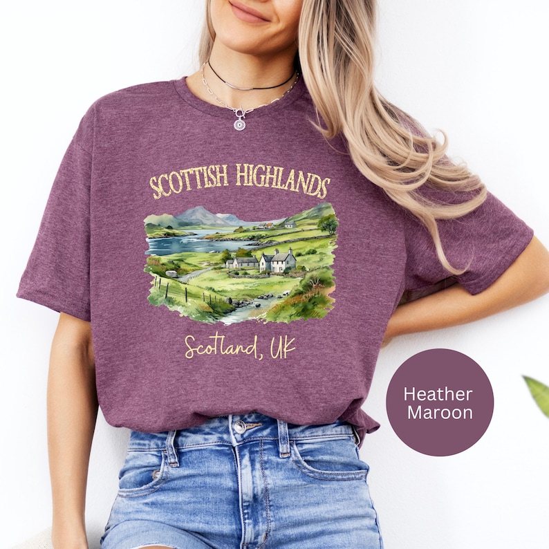Puede incluir: Camiseta color granate con una ilustraci&oacute;n en acuarela de las Tierras Altas escocesas. El dise&ntilde;o incluye un paisaje con monta&ntilde;as, un lago y casas, con el texto "Scottish Highlands" y "Scotland, UK".