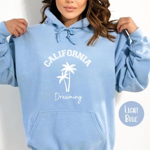 Puede incluir: Sudadera con capucha azul claro con un gr&aacute;fico blanco de palmeras y el texto "CALIFORNIA Dreaming".