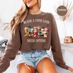 May include: A brown long-sleeve sweatshirt featuring the text "BECAUSE A COFFEE LOVER NEEDS OPTIONS" above and below a graphic illustration of various coffee drinks, mugs, and pastries including a croissant, muffin, and cookies.