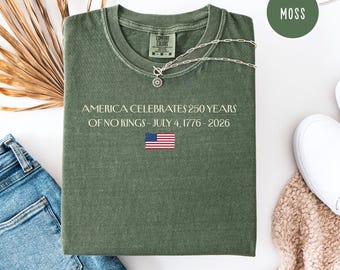 May include: A moss green t-shirt with the text "AMERICA CELEBRATES 250 YEARS OF NO KINGS - JULY 4, 1776 - 2026" and an American flag graphic. A silver necklace with a pendant is draped across the shirt. The shirt is folded on a white surface.
