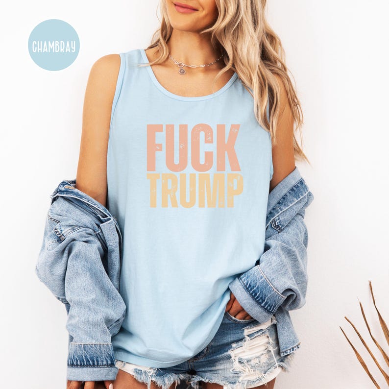 K&ouml;nnte beinhalten: Hellblaues Tanktop mit dem Text "FUCK TRUMP" in Pfirsich und Gelb. Das Tanktop ist &auml;rmellos und hat einen runden Ausschnitt. Eine Jeansjacke ist &uuml;ber den Schultern drapiert.