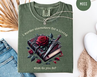May include: Moss green t-shirt with the text "i murder characters for a living, what do you do?" A graphic of a book, rose, and sword is printed on the front. A silver necklace is draped across the shirt.