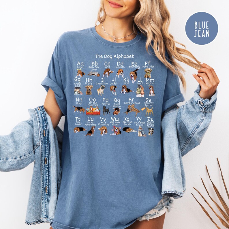May include: A blue jean t-shirt with a graphic of a dog alphabet. The alphabet is printed in white with a dog illustration for each letter. The text reads "The Dog Alphabet".