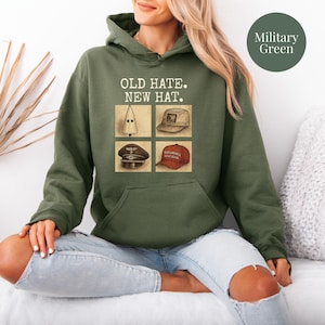 May include: Military green hoodie with the text "OLD HATE. NEW HAT." above four images of hats. The images include a white pointed hood, a tan cap, a black hat, and a red "MAKE AMERICA GREAT AGAIN" hat.