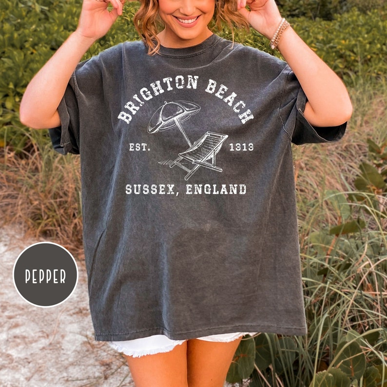 K&ouml;nnte beinhalten: Ein dunkelgraues T-Shirt mit einem wei&szlig;en Grafikdesign, auf dem "Brighton Beach Est. 1313 Sussex, England" steht. Die Grafik enth&auml;lt einen Sonnenschirm und einen Liegestuhl.
