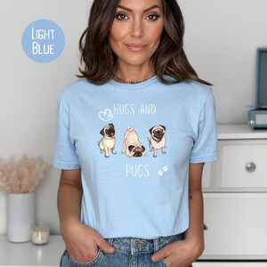 Puede incluir: Una camiseta azul claro con un gr&aacute;fico de tres carlinos y el texto "Hugs and Pugs".