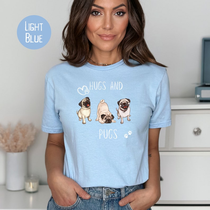 Puede incluir: Una camiseta azul claro con un gr&aacute;fico de tres carlinos y el texto "Hugs and Pugs".
