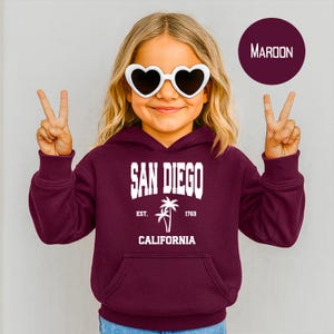 May include: A maroon hoodie with a front pocket and the words "SAN DIEGO CALIFORNIA" in white block letters. The hoodie features a palm tree graphic and the text "EST. 1769". The person is wearing heart-shaped sunglasses.