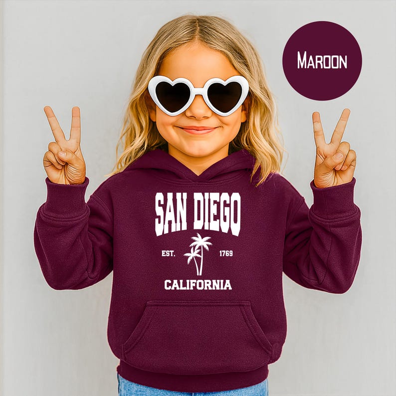 May include: A maroon hoodie with a front pocket and the words "SAN DIEGO CALIFORNIA" in white block letters. The hoodie features a palm tree graphic and the text "EST. 1769". The person is wearing heart-shaped sunglasses.