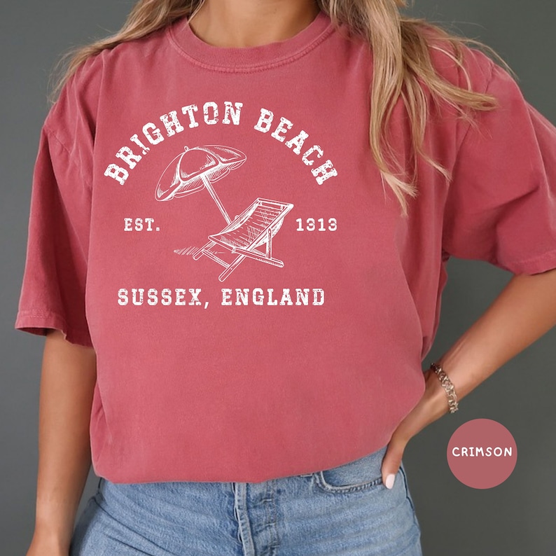 K&ouml;nnte beinhalten: T-Shirt in Rotbraun mit einem wei&szlig;en Grafikdesign, auf dem "Brighton Beach" mit einer Illustration von Sonnenschirm und Strandliege steht. Der Text lautet au&szlig;erdem "Est. 1913" und "Sussex, England".