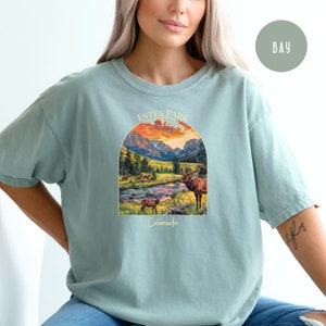 May include: A light blue t-shirt featuring a graphic print of a mountain scene with a sunset and elk. The text "Estes Park Colorado" is printed below the graphic.